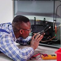 Young Male Serviceman Working On Fridge With Screwdriver At Home
